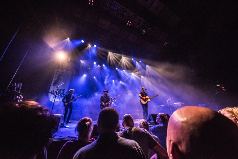 Een liveband speelt op een podium met felgekleurde lichten, terwijl het publiek naar de voorstelling kijkt.