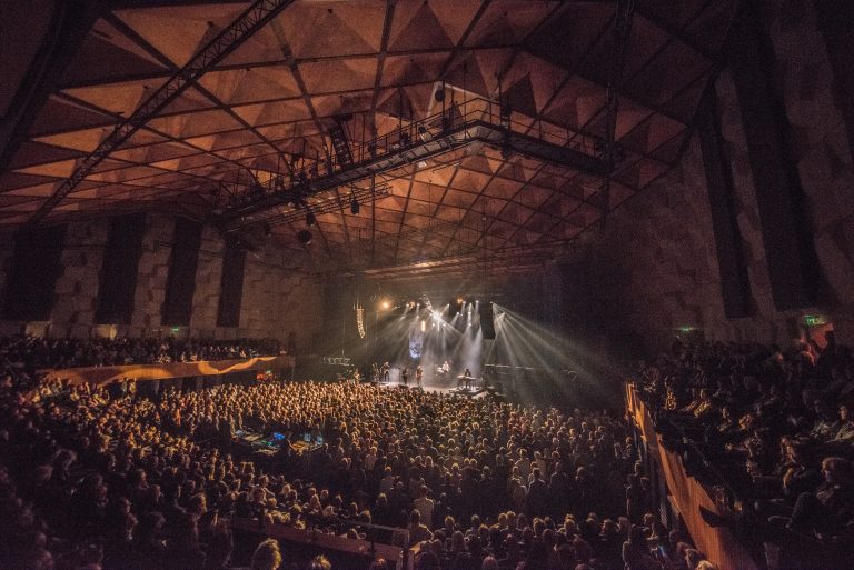Een drukbezocht concert in een grote, moderne zaal met sfeervolle verlichting.
