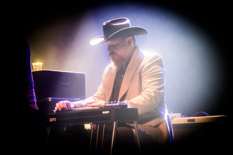 Een muzikant in een hoed speelt op een steelgitaar in een sfeervolle, verlichte setting.