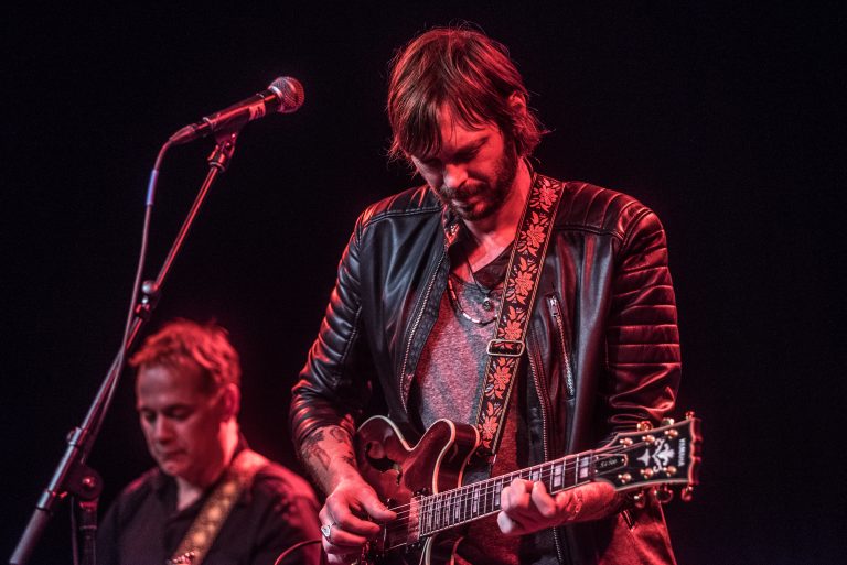Een muzikant speelt gitaar op een podium, terwijl een andere muzikant op de achtergrond te zien is.