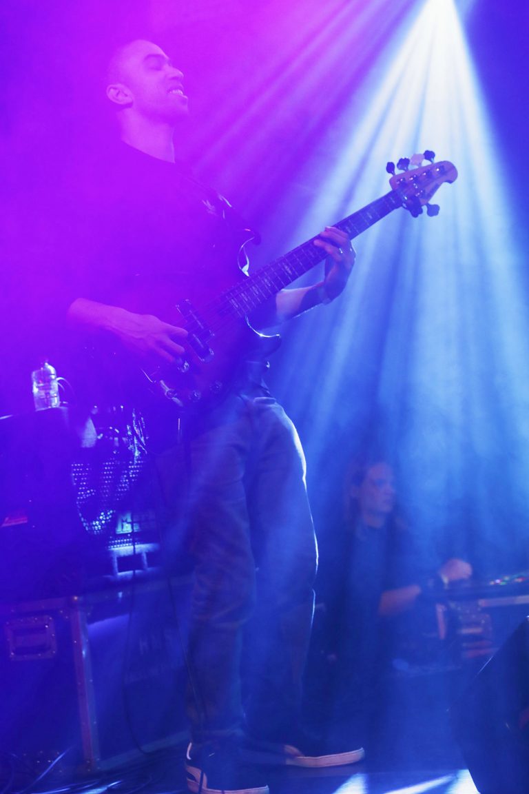 Een muzikant speelt een elektrische basgitaar onder kleurrijke lichteffecten op een podium.