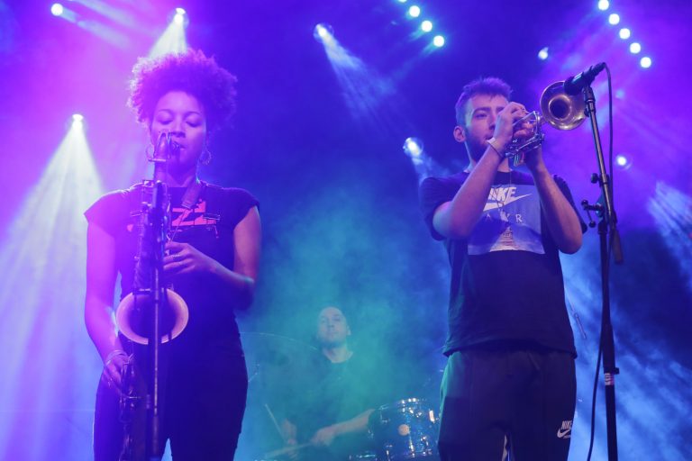 Een zangeres met een saxofon en een trompettist in een kleurrijke, verlichte muzieksetting.