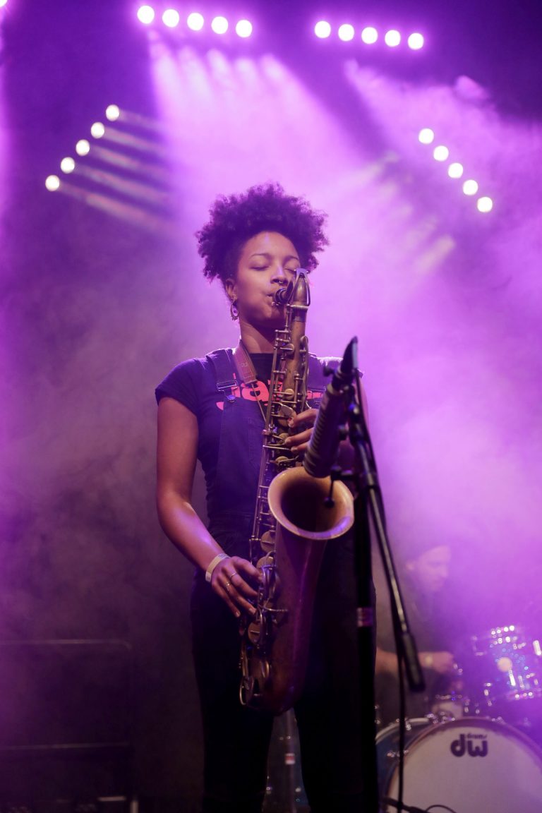 Een muzikante met een saxofoon staat op het podium in een paarse lichtshow met rook.