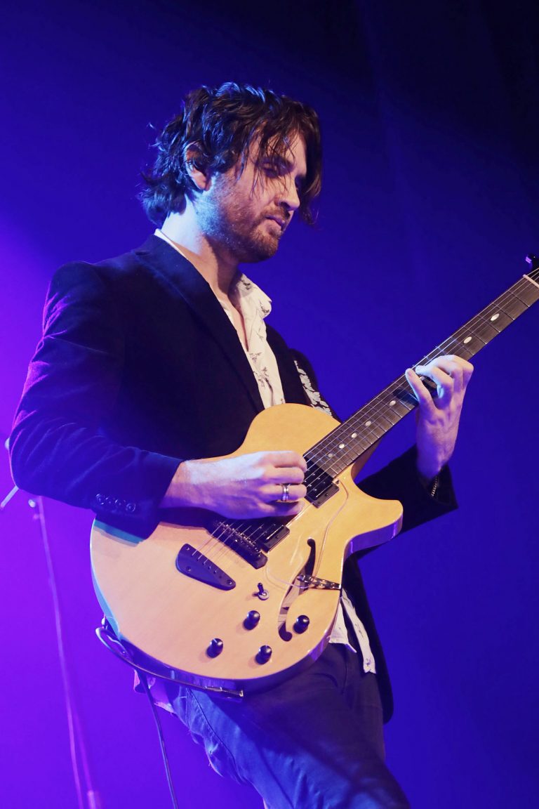 Een muzikant speelt gitaar op het podium met paarse belichting.