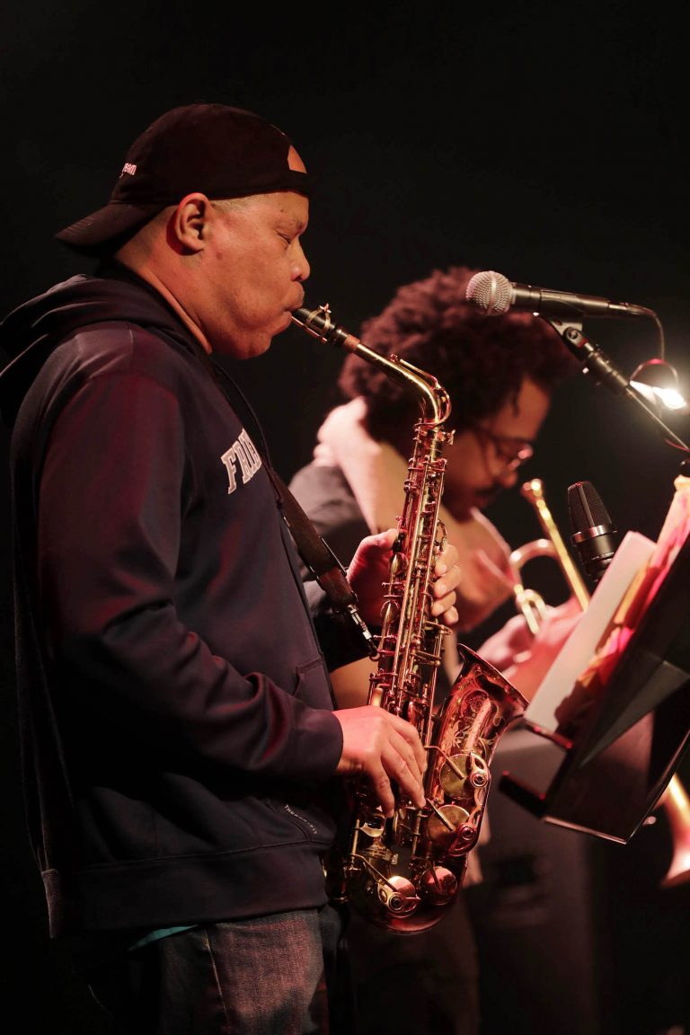Twee muzikanten spelen op een podium, één bespeelt een saxofoon en de ander een trompet.