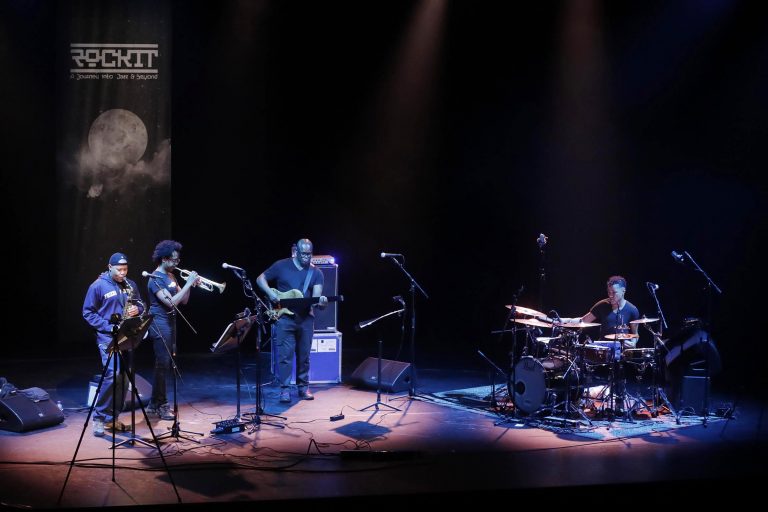 Een muzikale uitvoering op een podium met drie artiesten die verschillende instrumenten bespelen.