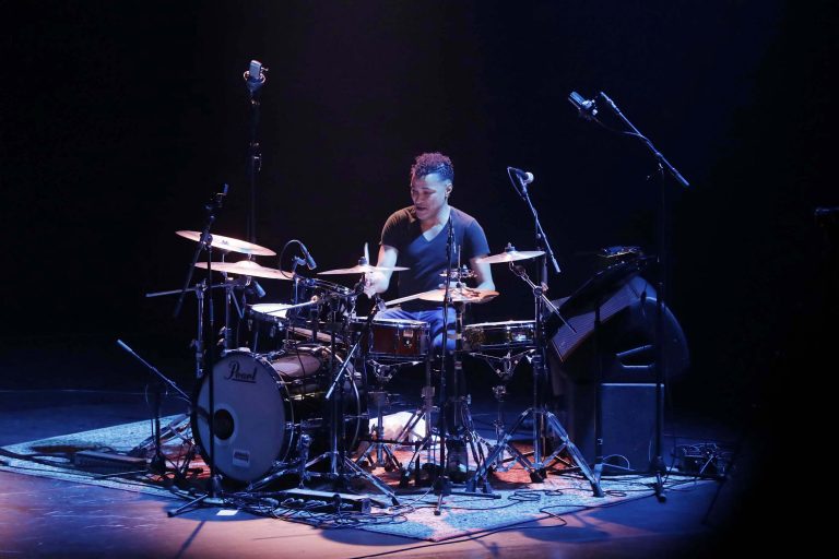 Een drummer die op een set drums speelt, verlicht door blauwe en paarse lichten in een donkere omgeving.