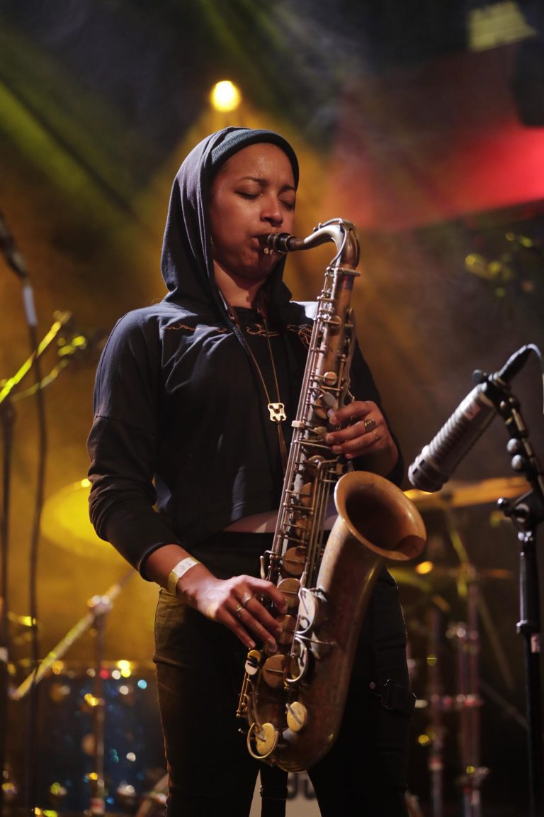 Een muzikante speelt saxofon op een podium, omringd door kleurrijke verlichting.