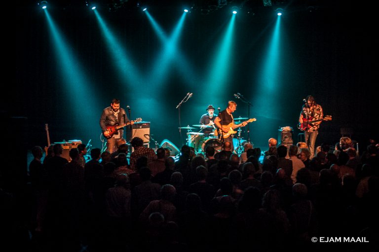 Een band speelt live op het podium terwijl het publiek geniet van de show, omgeven door sfeervolle blauwe verlichting.
