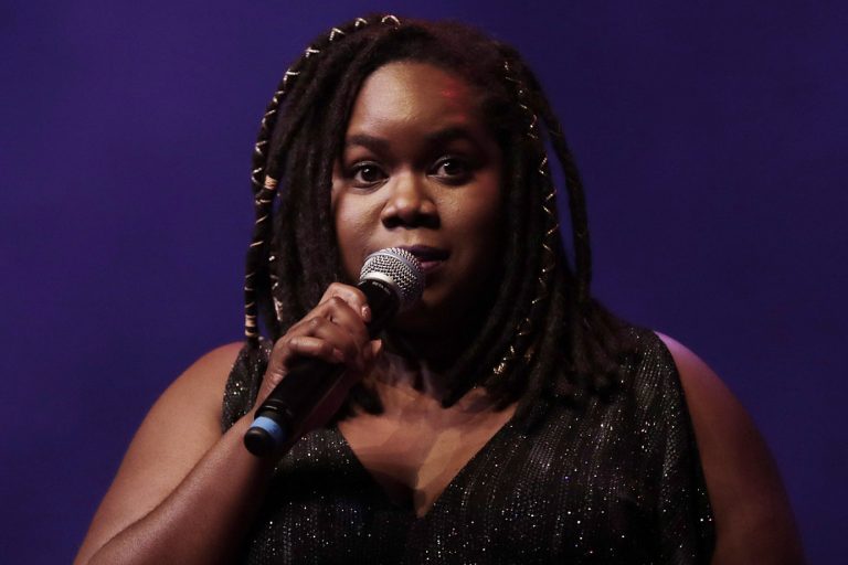 Een vrouw met dreadlocks houdt een microfoon vast en kijkt recht in de camera, terwijl ze zich op een podium bevindt.