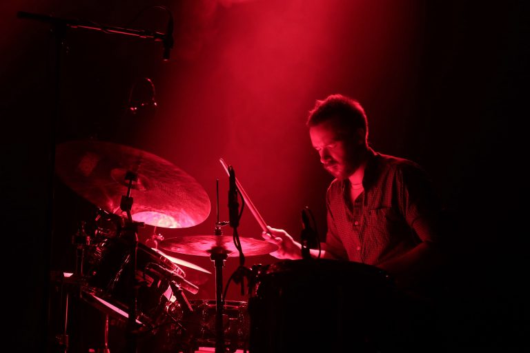 Een drummer speelt energiek op zijn drums onder een rode gloed van licht.