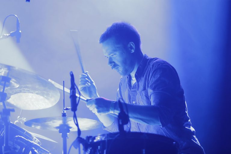 Een drummer die gefocust speelt op zijn drums, omringd door blauw licht.