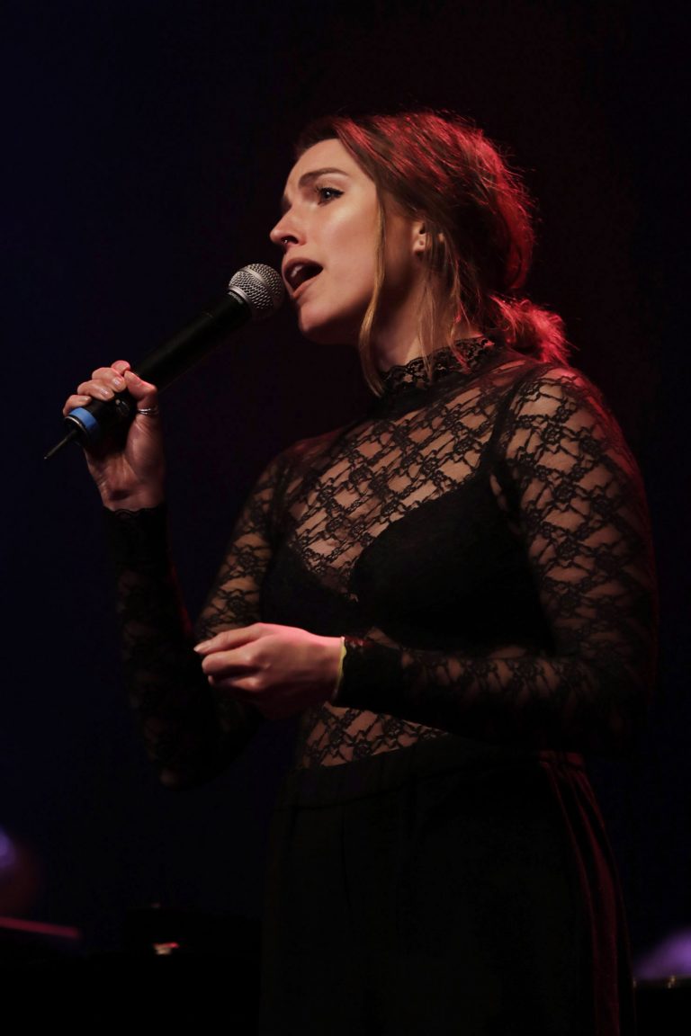 Een vrouw in een zwart doorschijnend shirt zingt met een microfoon op een podium.