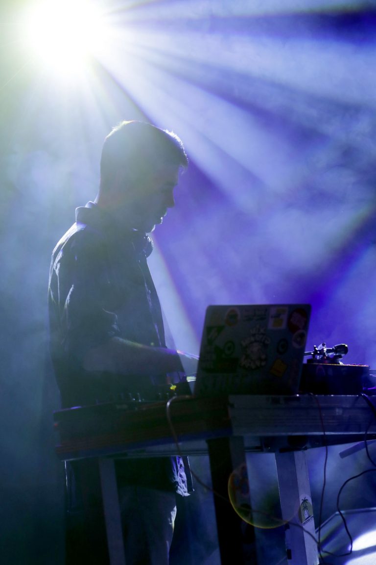 Silhouet van een DJ die aan een laptop werkt, omringd door paarsachtig licht en mist.