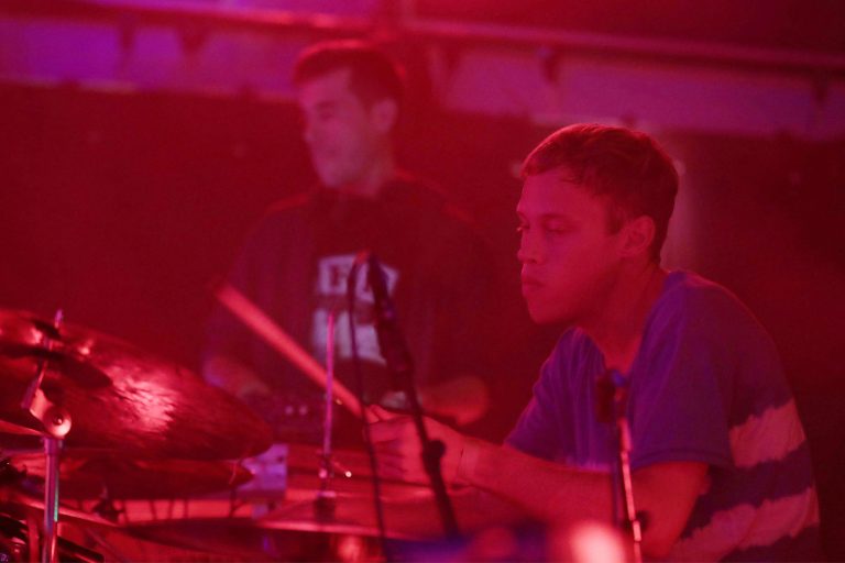 Twee muzikanten spelen op een podium met rode verlichting, waarbij de focus ligt op de drummer.