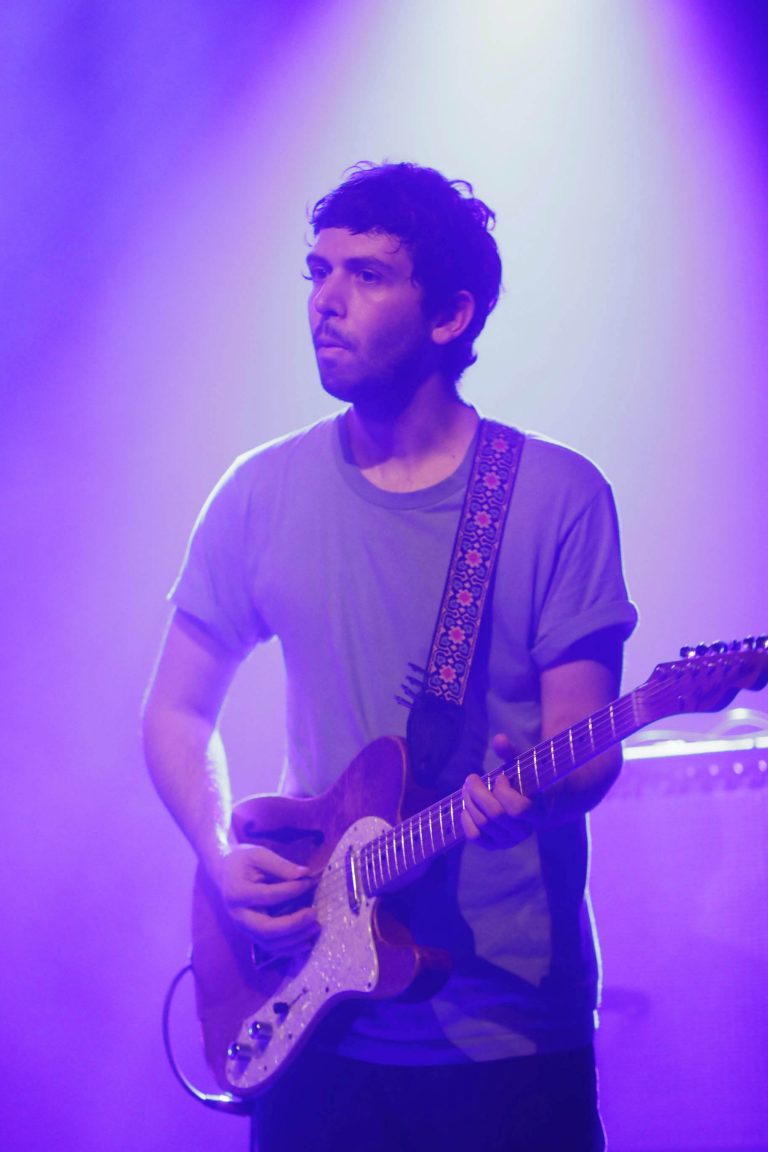 Een muzikant speelt gitaar op een podium met paarse belichting.