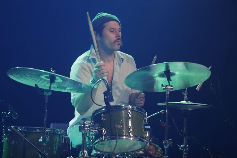 Een drummer speelt op een set met cymbals en een snaredrum, verlicht door een blauwe gloed.