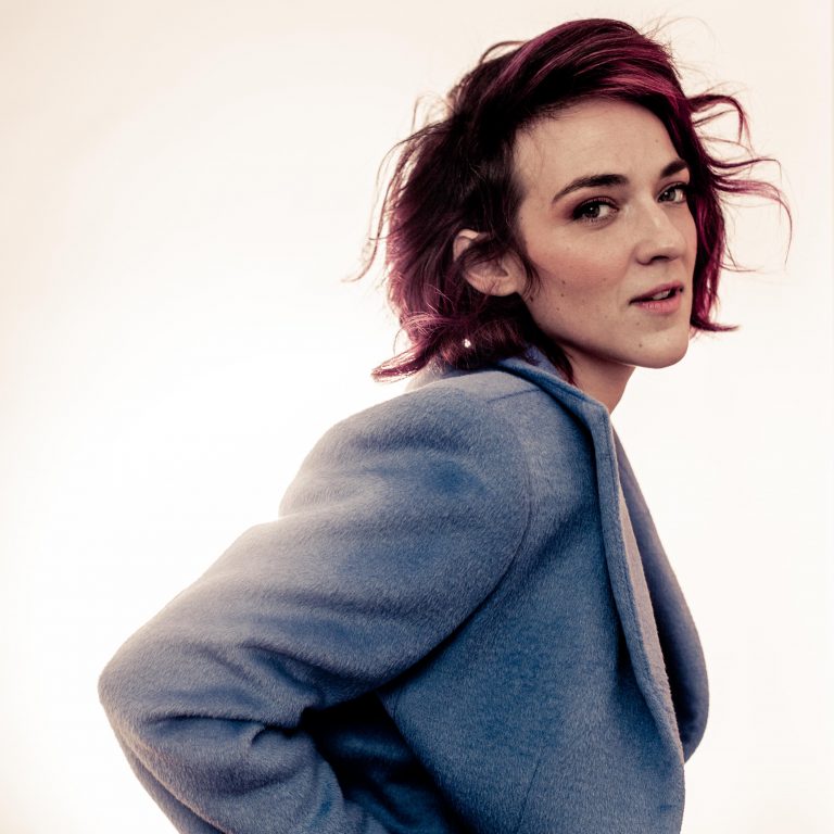 Een persoon met een schouderlange, paarsachtige haarkleur kijkt over de schouder, gekleed in een blauwe blazer tegen een lichte achtergrond.