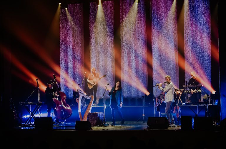 Een live muziekoptreden met verschillende muzikanten op het podium, omringd door kleurrijke lichtstralen.
