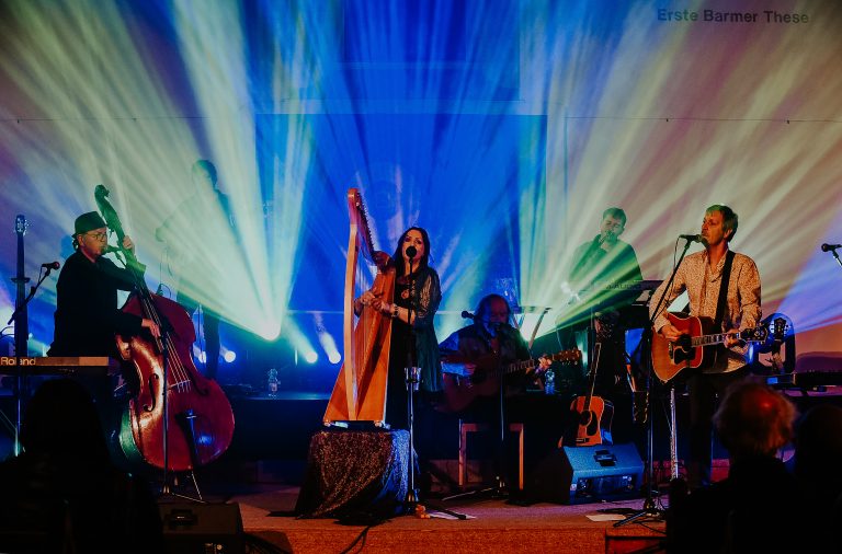 Een muzikante met een harp, omringd door andere instrumentalisten, staat op het podium onder kleurrijke lichten.