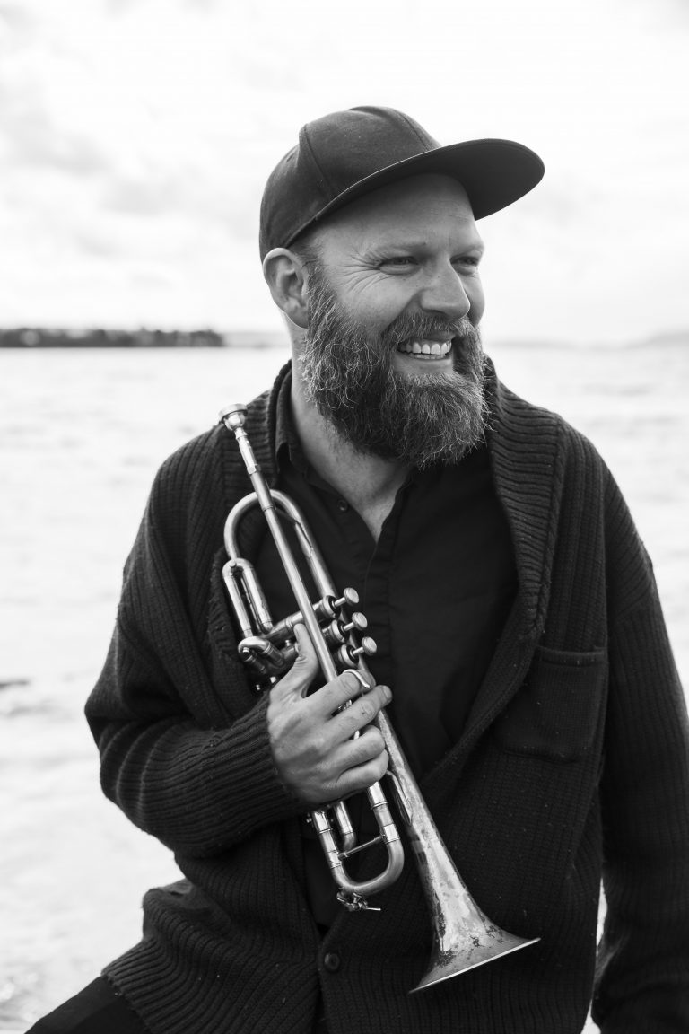 Een man met een baard en een pet houdt een trompet vast, met water op de achtergrond.