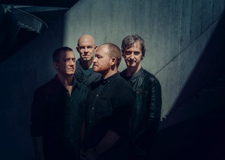 Vier mannen in zwarte kleding poseren tegen een betonnen muur met schaduwen die een dramatisch effect creëren.