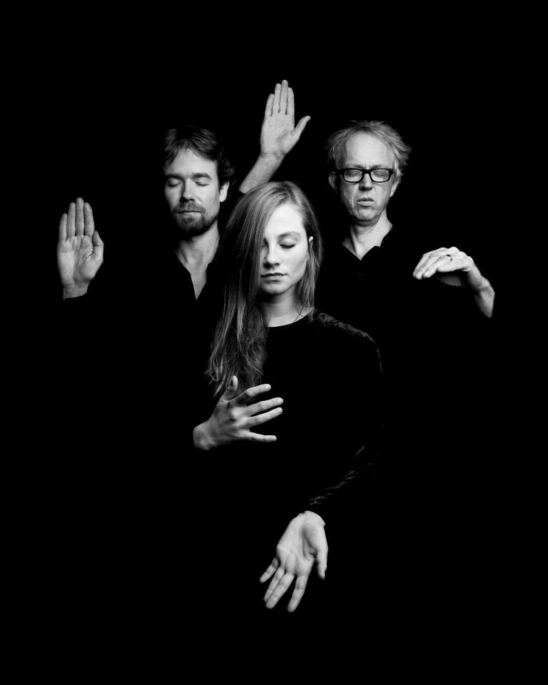 Drie mensen in zwarte kleding, met verschillende handhoudingen, tegen een zwarte achtergrond.