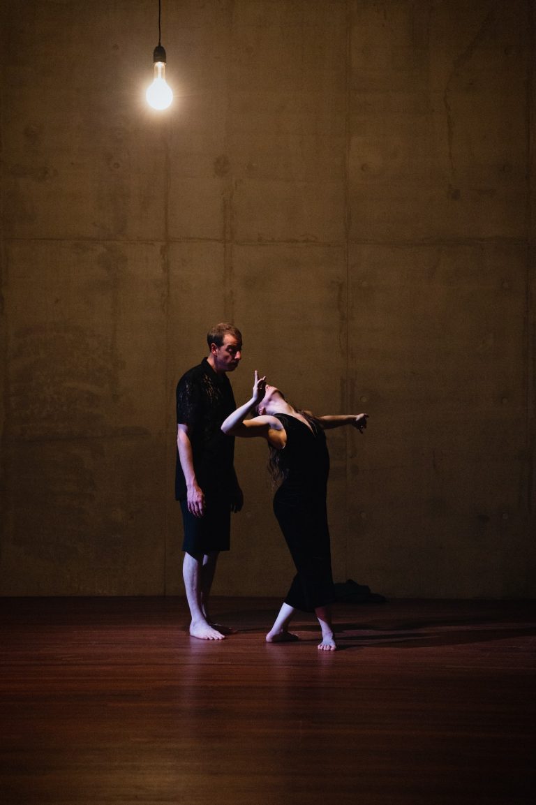Twee dansers, een man en een vrouw, in een intieme pose tegen een eenvoudige, betonnen achtergrond.
