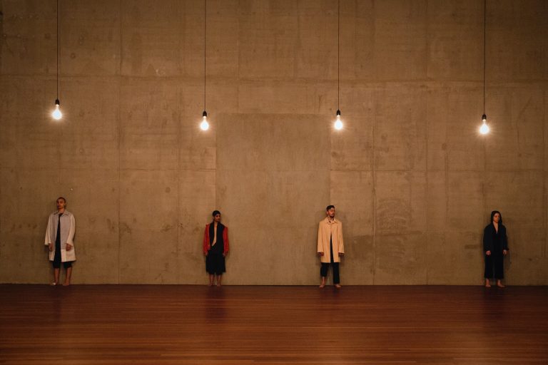 Twee mensen staan naast elkaar tegen een betonnen muur, met hangende lampen boven hen.