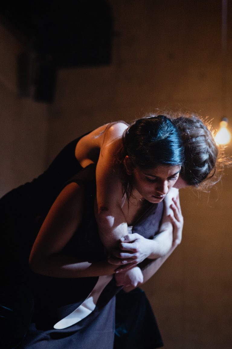 Twee dansers zijn in een intieme pose, waarbij één danser de ander vasthoudt in een donkere setting met sfeerverlichting.