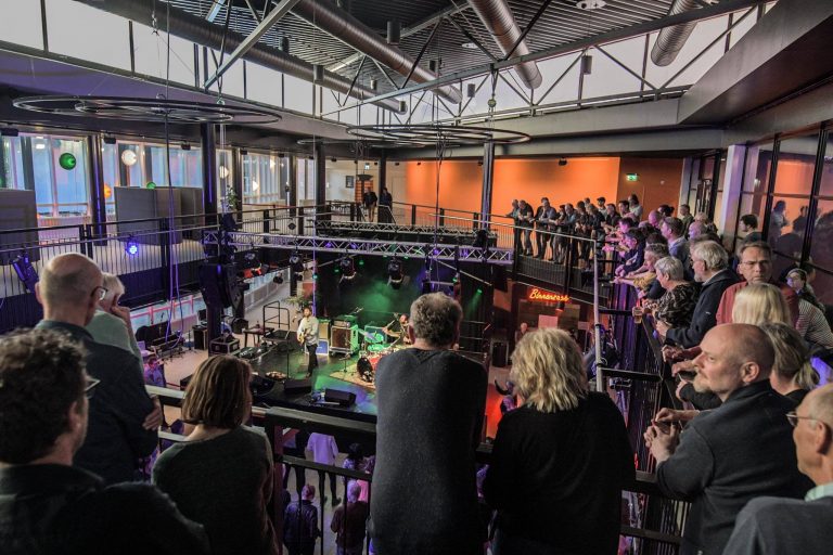 Een live muziekoptreden met een menigte die van bovenaf toekijkt in een moderne, industriële ruimte.