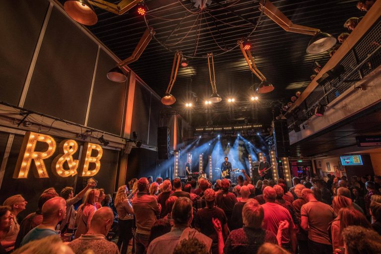 Een levendige concertzaal met een enthousiast publiek dat naar een optreden kijkt, omgeven door kleurrijke verlichting en decoraties.