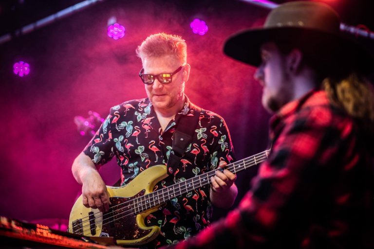 Een muzikant speelt basgitaar op het podium, omgeven door kleurrijke verlichting en rook.