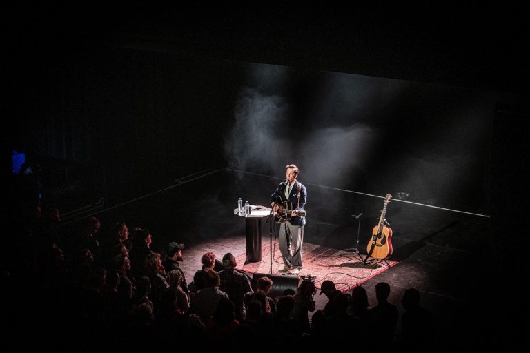 Een muzikant staat op een podium met een gitaar, terwijl het publiek naar de voorstelling kijkt in een sfeervolle, rookachtige setting.