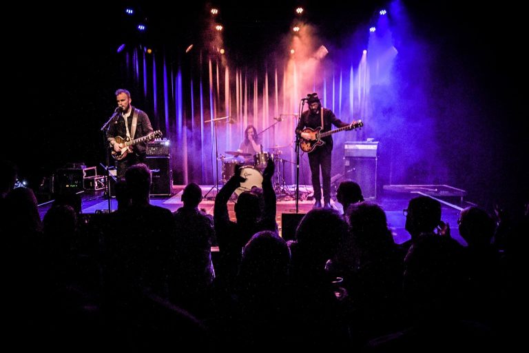 Een band speelt live muziek op een podium met kleurrijke verlichting en een enthousiast publiek ervoor.