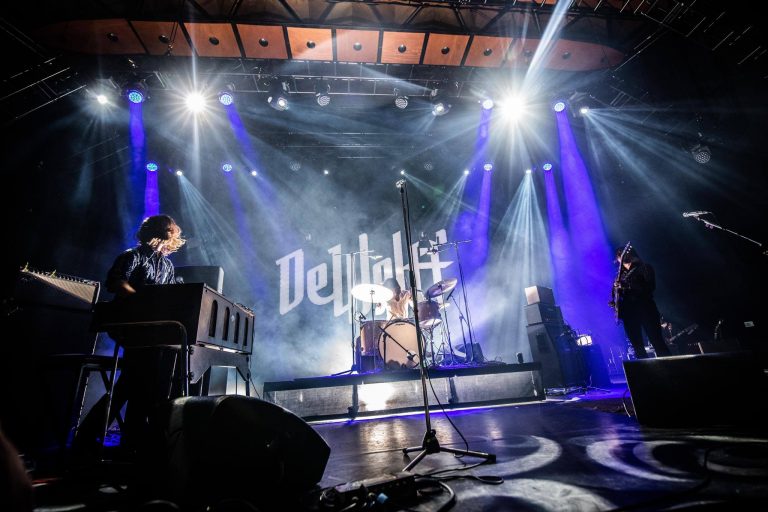 Een muzikale optreden met een band op een podium, omgeven door kleurrijke verlichting en een projectie van de bandnaam op de achtergrond.