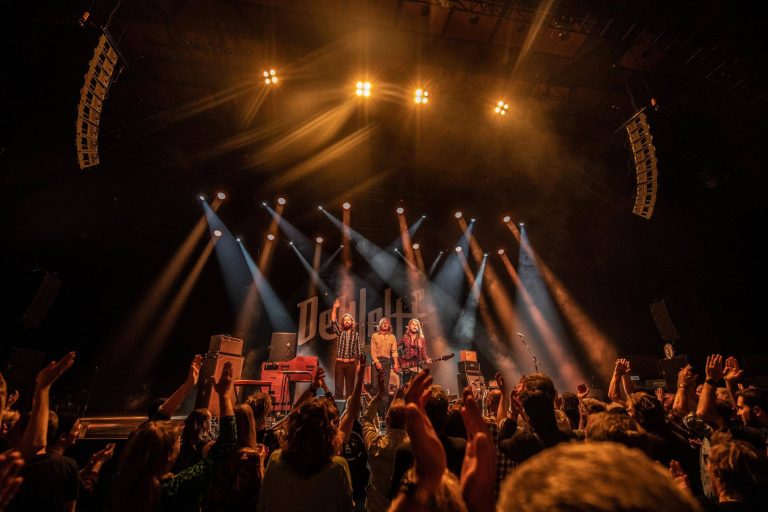 Een levendig concert met een band op het podium en een enthousiast publiek dat zijn handen in de lucht steekt.