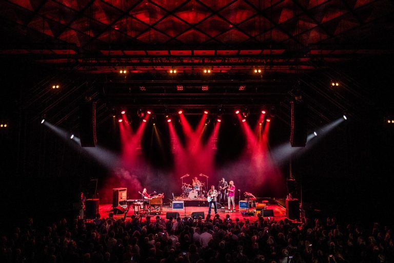 Een band optredend op een podium, verlicht door felrode lichten, met een groot publiek ervoor.
