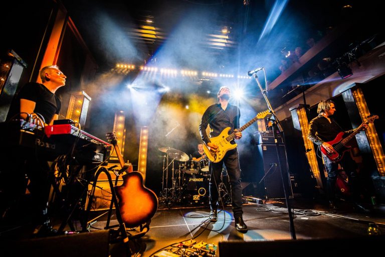 Een muzikant staat op een podium met een gitaar, omringd door sfeervolle verlichting en rook.
