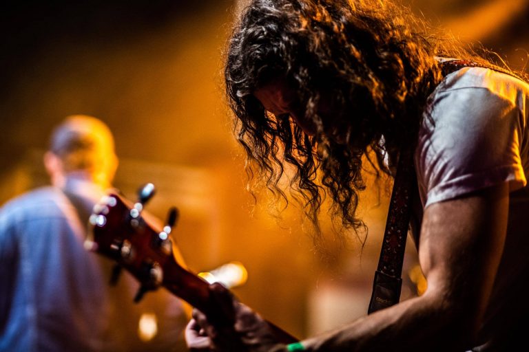 Een muzikant met krullend haar speelt gitaar op het podium, omgeven door een warme gloed van licht.