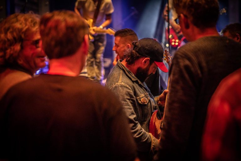 Een man in een spijkerjack speelt een gitaar voor een publiek op een podium met kleurrijke verlichting.