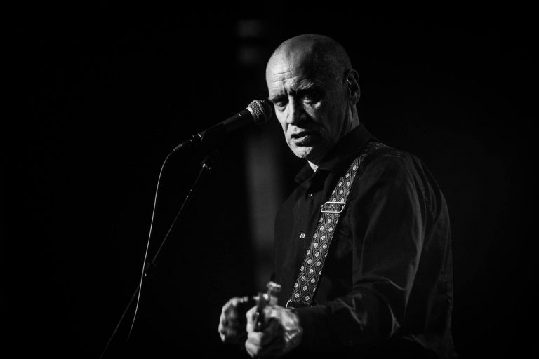 Zwart-witfoto van een muzikant die een gitaar speelt en in de richting van de camera kijkt.