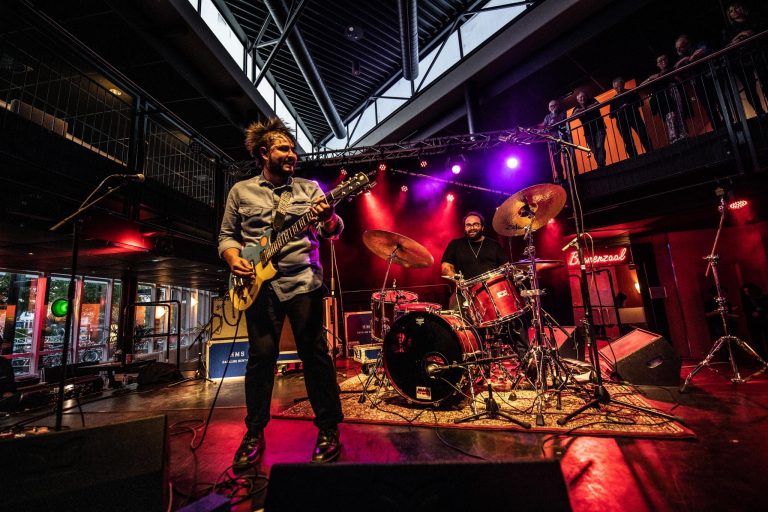Een band speelt op een podium met kleurrijke verlichting, waarbij de gitarist en drummer zichtbaar zijn.