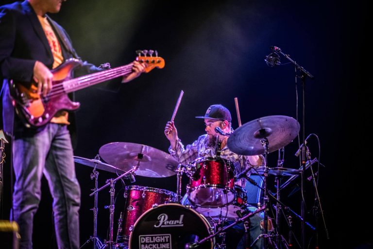 Een drummer speelt op een rode drumset terwijl een bassist in de achtergrond staat tijdens een optreden.