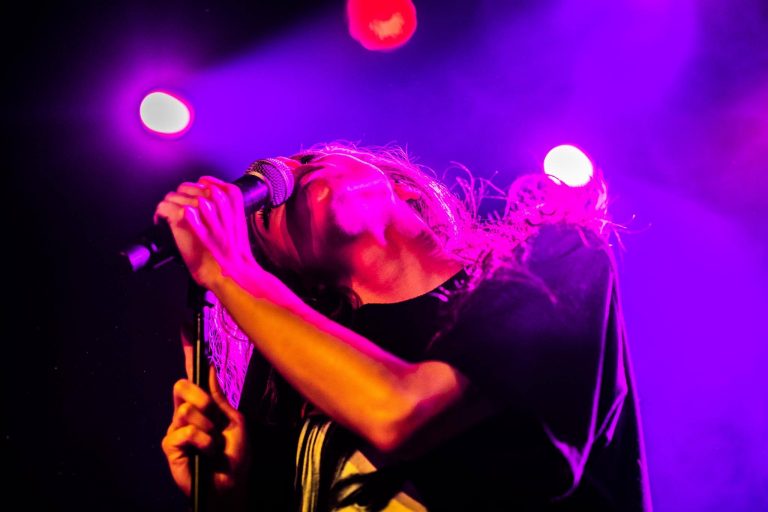 Een muzikant die intens in het optreden opgaat, met een microfoon in de hand en onder kleurrijk licht.