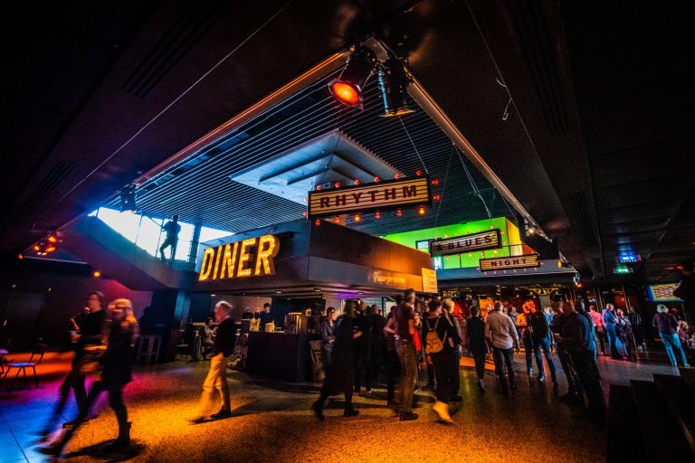Een drukke diner-ruimte met felgekleurde verlichting en mensen die zich verzamelen rondom een bar.