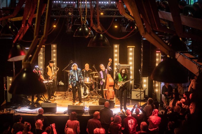 Een band speelt live op een podium met kleurrijke lichten, terwijl het publiek naar hen kijkt.