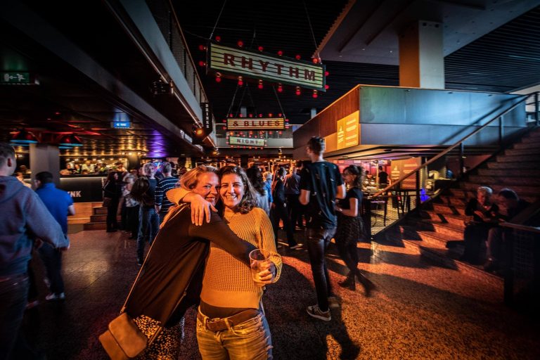 Twee vrouwen poseren vriendelijk voor de camera in een levendige omgeving vol mensen en kleurrijke verlichting.