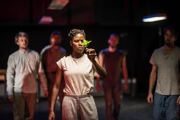 Een vrouw houdt een groene vlinder voor haar gezicht terwijl ze met anderen in een donkere ruimte staan.