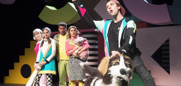 Een groep performers op een kleurrig podium met een man die een hond vasthoudt en enthousiast spreekt.